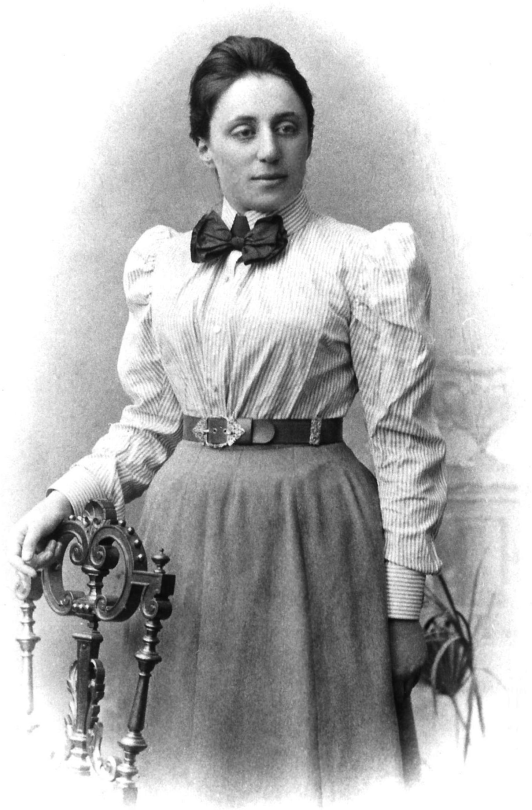 A greyscale photo of mathematician Emmy Noether. She is wearing a puffy pinstriped shirt, a long grey skirt, and a bow tie.